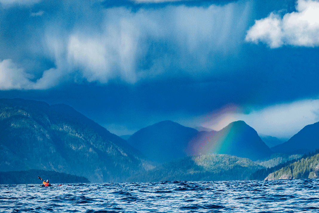 a long distance expedition kayaker paddles the cloudy Kyuquot Crossing in front of a rainbow