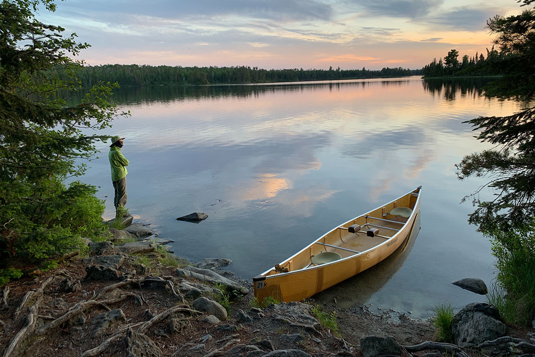 man stands on shore beside a beached yellow canoe at dawn
