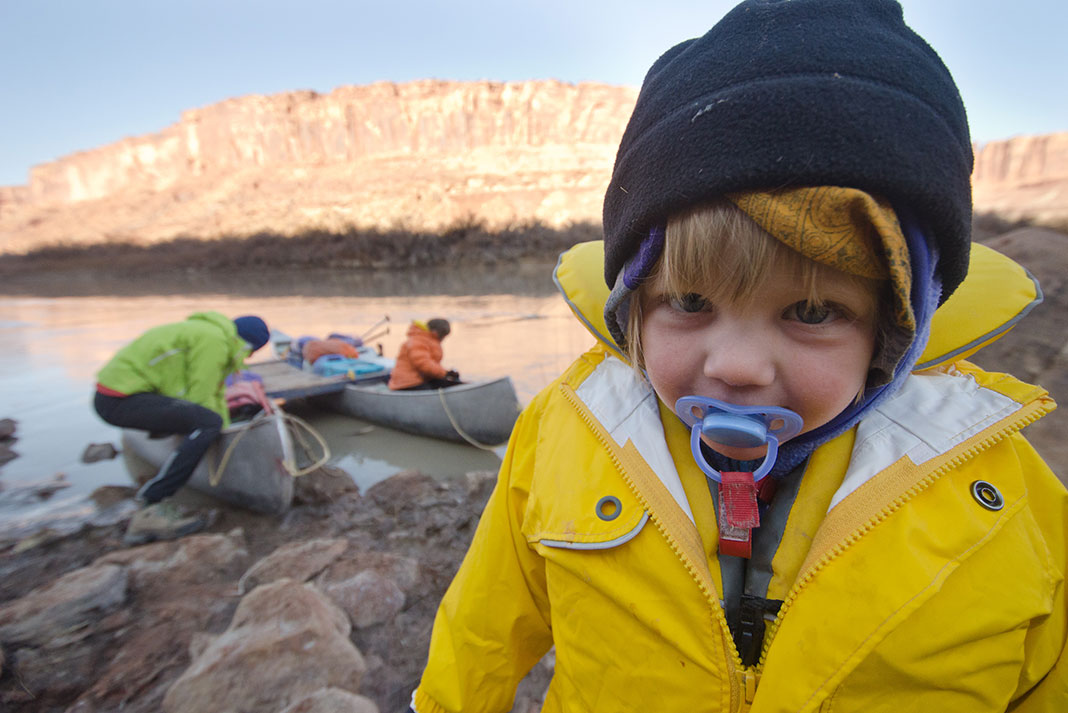 young child with soother and rain coat stands in front of family preparing canoes on a camping trip