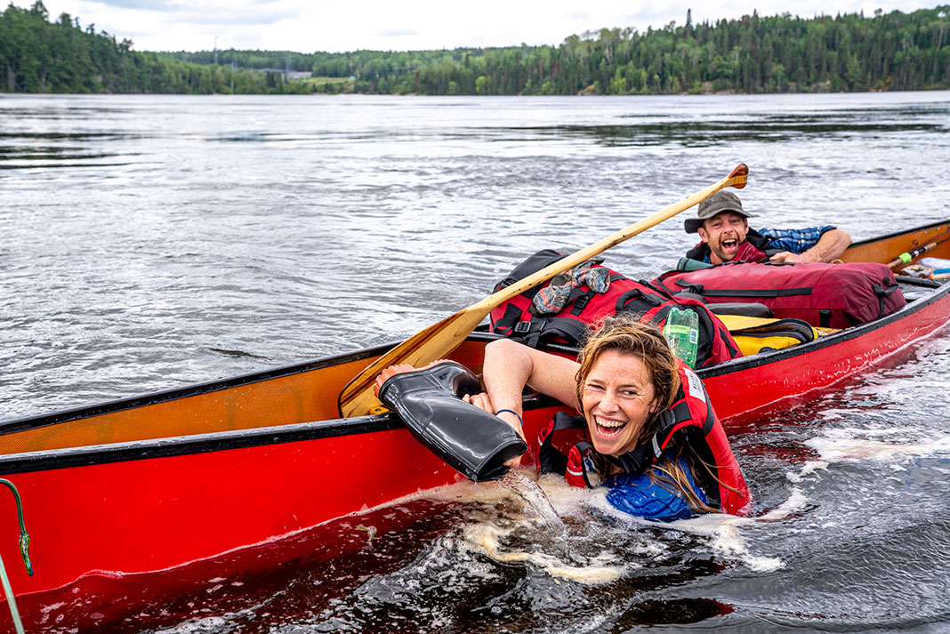 man and woman swim beside swamped canoe while emptying water out of rubber boots
