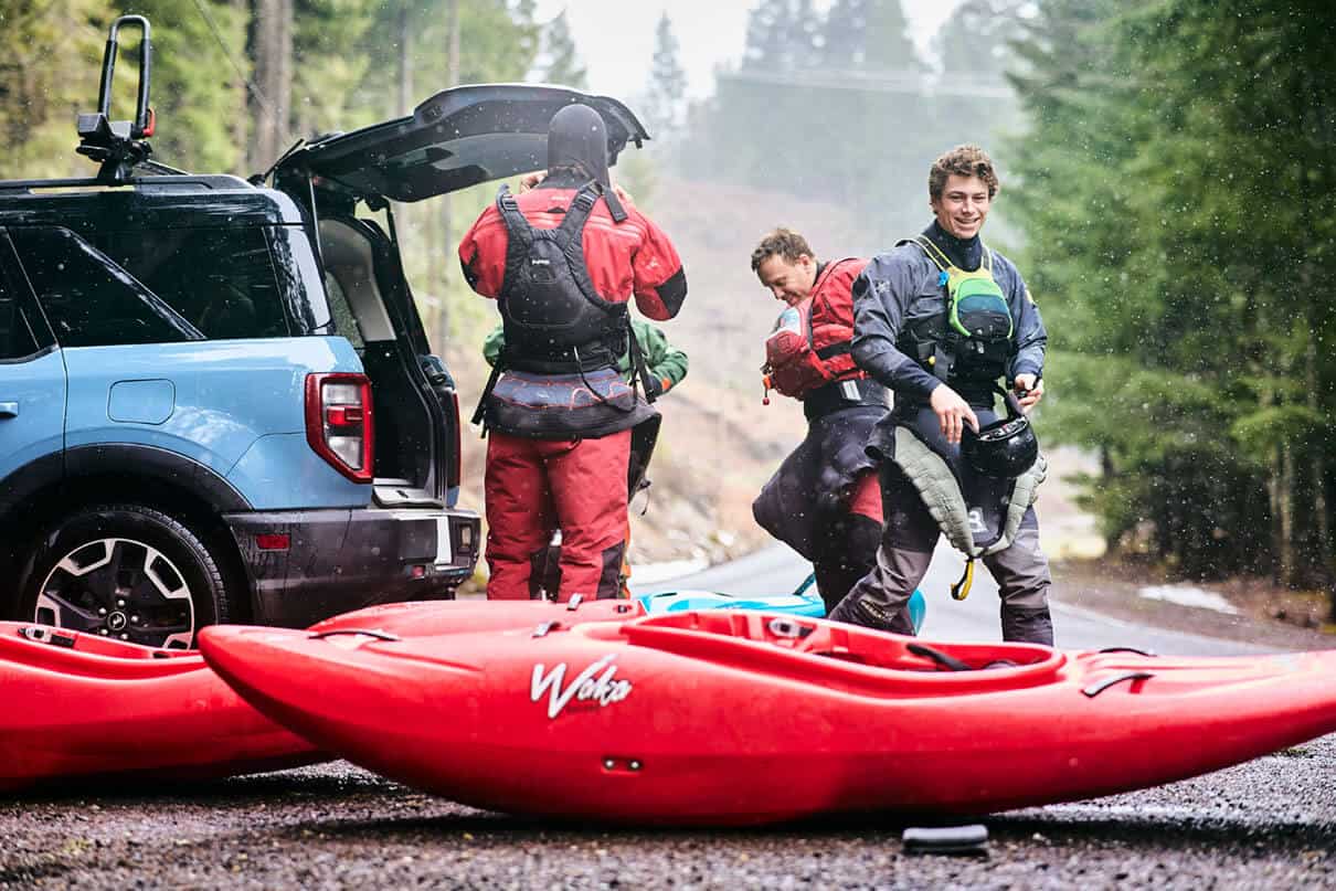 three whitewater kayakers gear up by the trunk of their vehicle with red kayaks on the ground in front