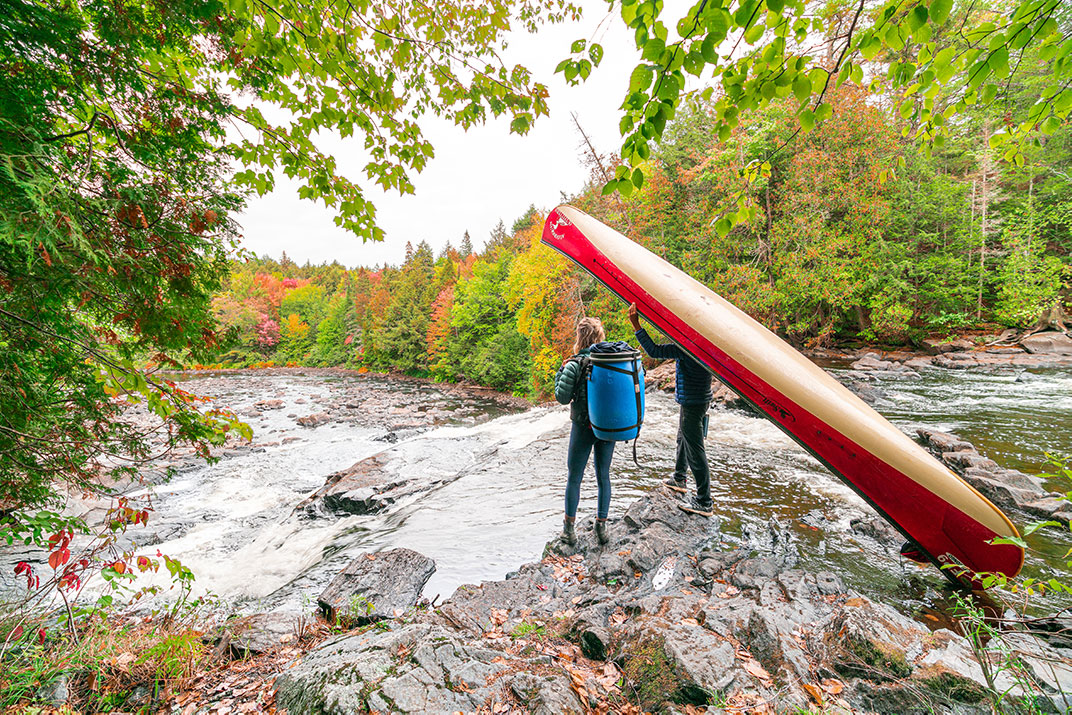 Two people standing at top of waterfall, one with canoe and other with blue barrel.