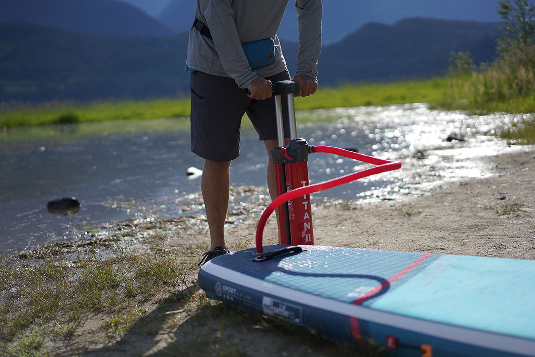 a person pumps up an inflatable paddleboard in front of water and hills