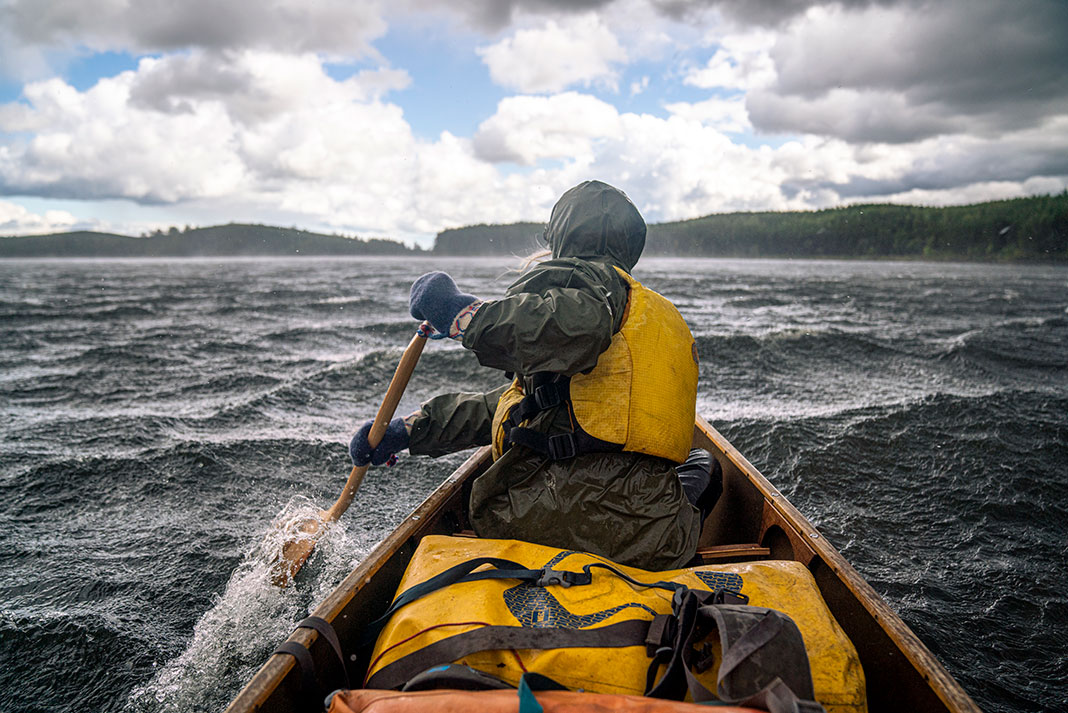 person in rain gear paddles a canoe across a choppy lake