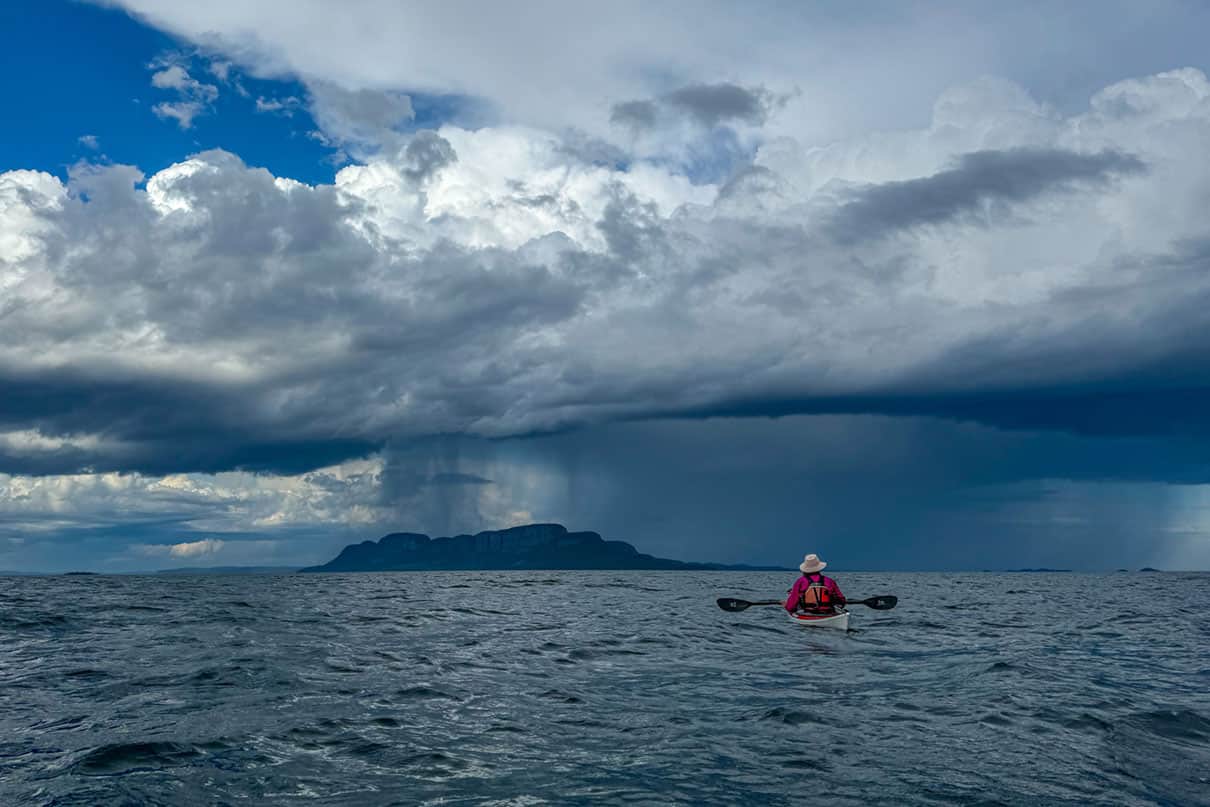 A sea kayaker sits on the water looking at a storm on the horizon