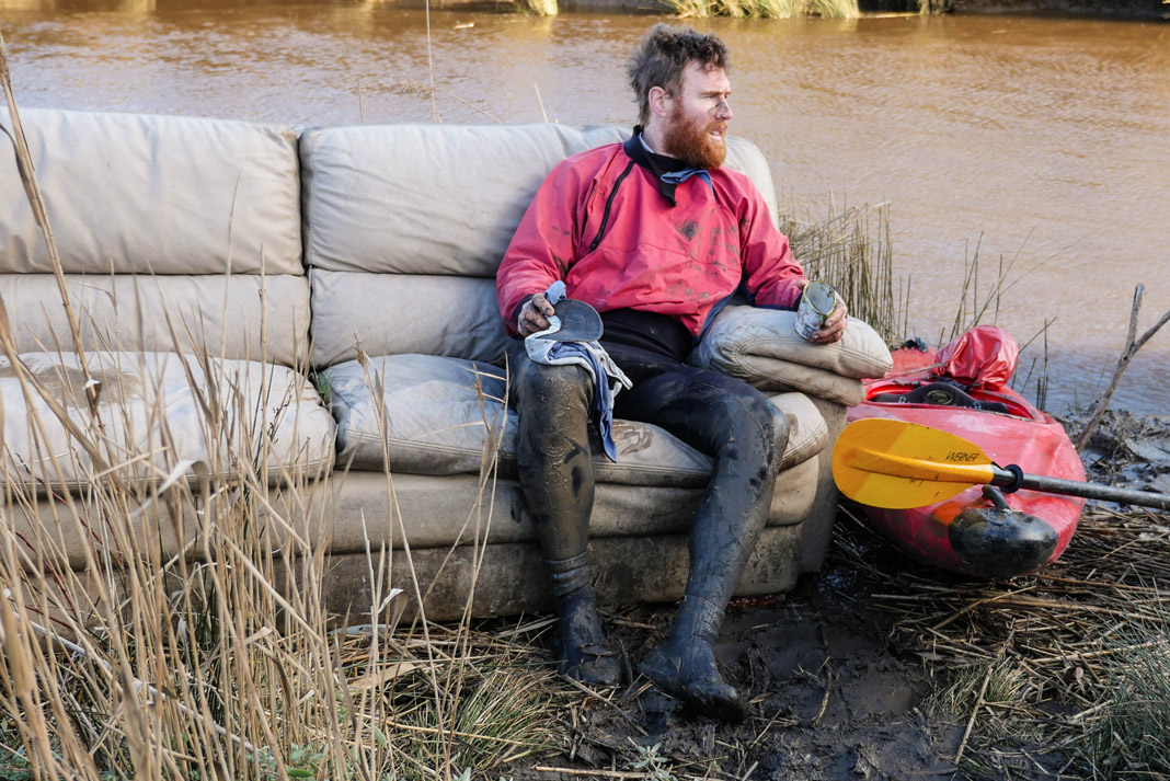 Beau Miles sitting on a couch next to his kayak and his kayak gear