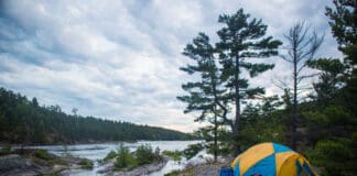 a yellow and blue tent sits on a rocky canoe campsite beside the water