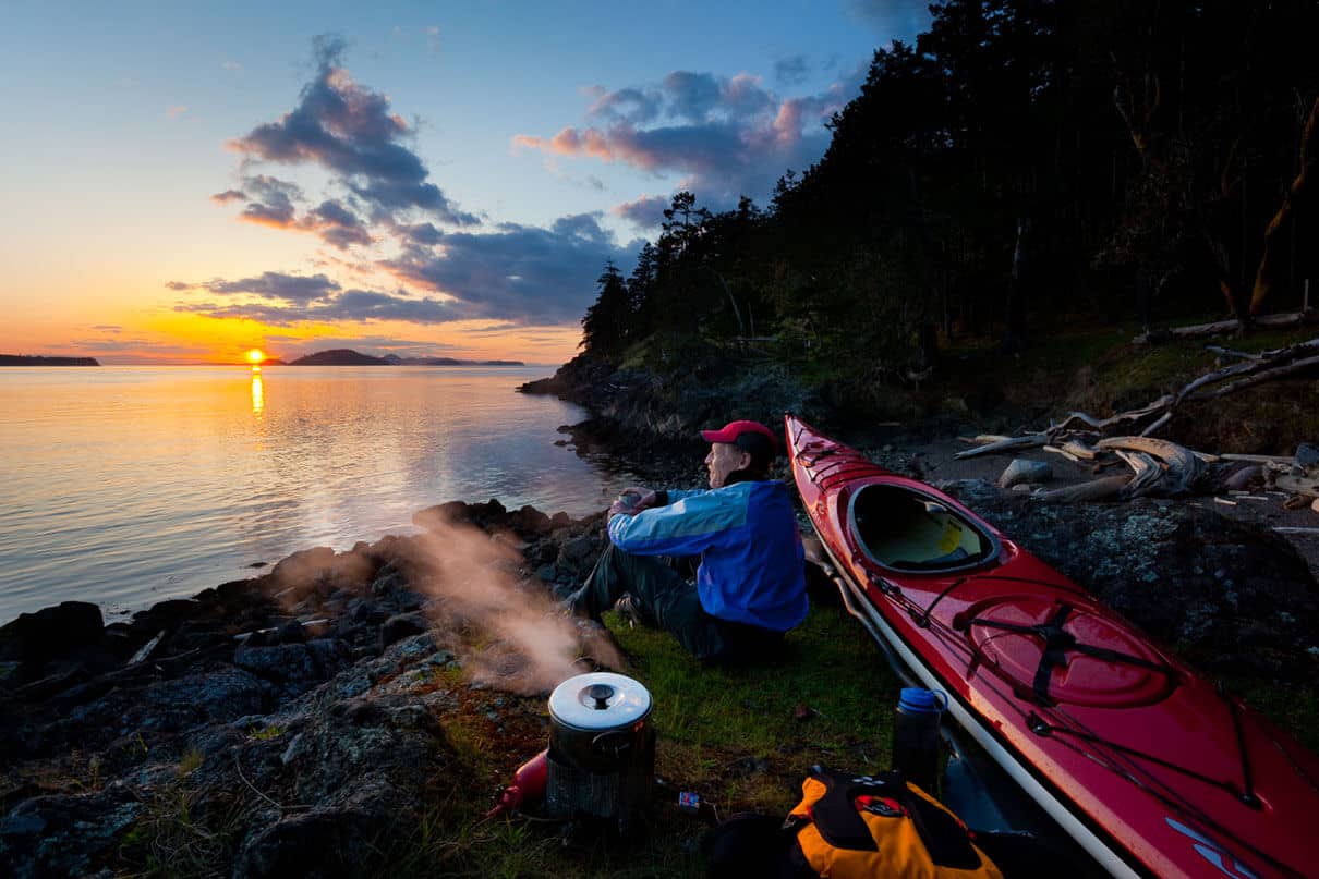 man sits beside his sea kayak at a campsite at dawn