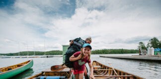 girl smiles and looks into the camera as she wades between two canoes while carrying a canoe pack at an outdoor education program, with instructor behind her