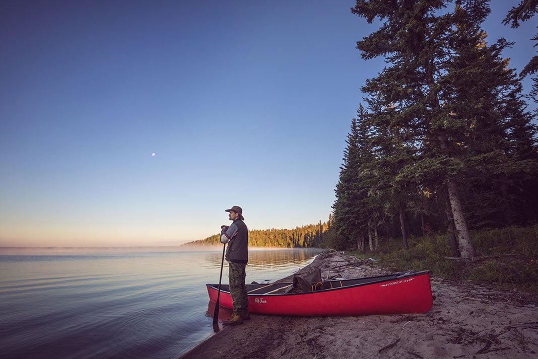 man stands at lake's edge holding a paddle beside a tripping canoe at dawn