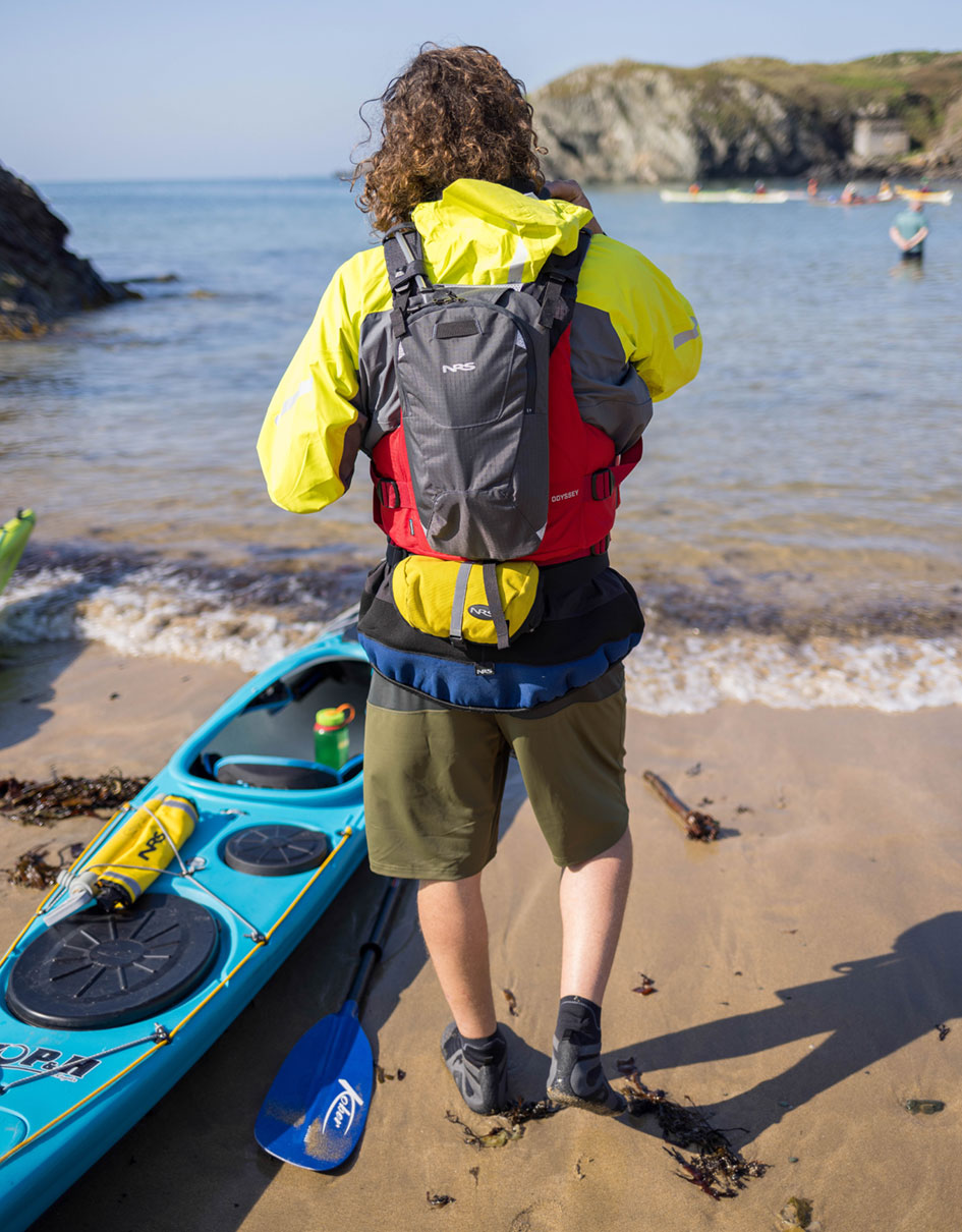Person wearing NRS Wetshoe while standing on a sand beach beside a kayak.